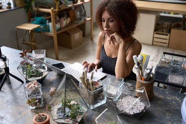 美しいブルネットの女性はノートパソコンの画面を見ながらおいしい装飾された植物ワークショップに座って - 写真・画像