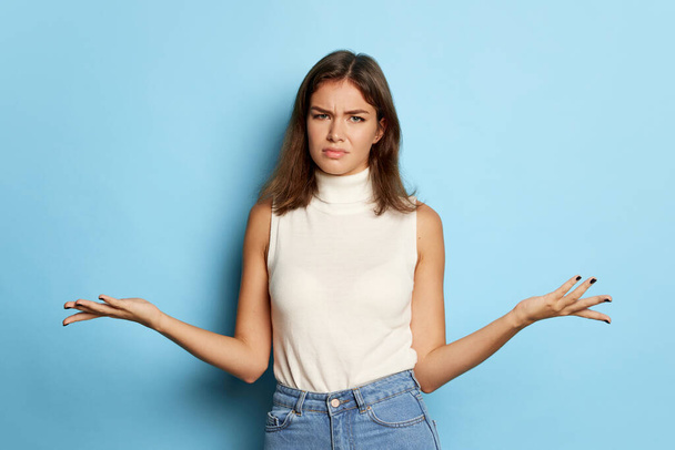 Portrait of young beautiful woman posing with asking expression isolated over blue background. Need in explanation. Concept of youth culture, emotions, facial expression, fashion. Copy space for ad - 写真・画像