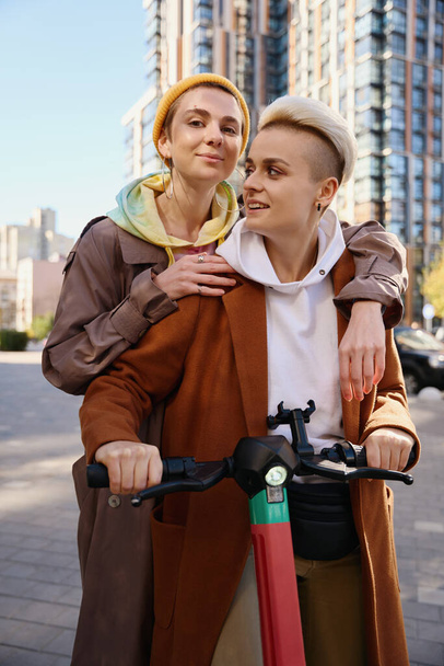 Blonde and brunette ride together on scooter around the city on an autumn sunny day, against the backdrop of skyscrapers - 写真・画像