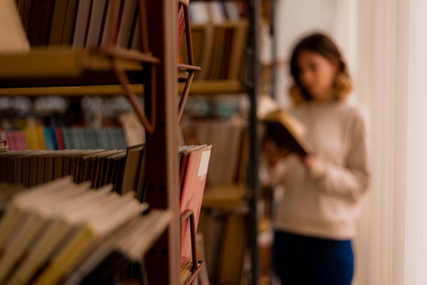Blurred college girl reading a book while standing besides the bookshelves in the library - Фото, зображення