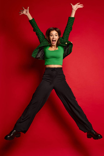 Portrait of young beautiful woman posing, jumping in excitement isolated over red studio background. Green outfit. Concept of youth culture, emotions, facial expression, fashion. Copy space for ad - Photo, image