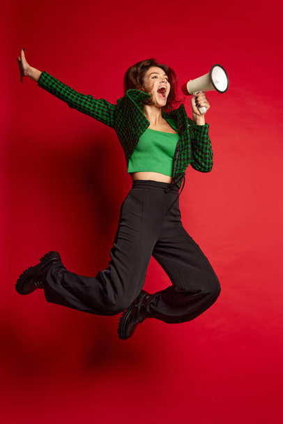 Portrait of young beautiful woman posing, shouting in megaphone in a jump isolated over red background. Holiday sales. Concept of youth culture, emotions, facial expression, fashion. Copy space for ad - Photo, image