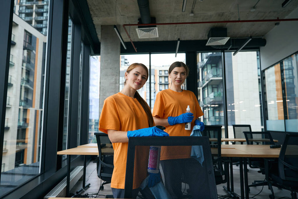 Young women are in the office area in work clothes and rubber gloves, they have wipes and cleaning products - Fotoğraf, Görsel