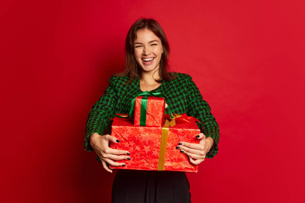 Portrait of young beautiful woman posing in happiness with holiday present boxes isolated over red studio background. Concept of youth culture, emotions, facial expression, fashion. Copy space for ad - Fotoğraf, Görsel