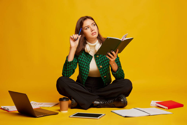 Portrait of young beautiful woman, student sitting on floor and doing homework, posing isolated over yellow studio background. Concept of youth culture, emotions, facial expression, fashion, education - Fotografie, Obrázek