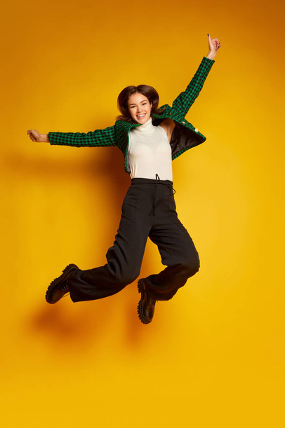 Portrait of young beautiful woman posing, cheerfully jumping isolated over yellow studio background. Concept of youth culture, emotions, facial expression, fashion. Copy space for ad - Φωτογραφία, εικόνα