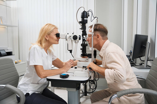 Doctor in white coat sitting at the table opposite the patient and using slit lamp to perform cycloscopy - Fotografie, Obrázek