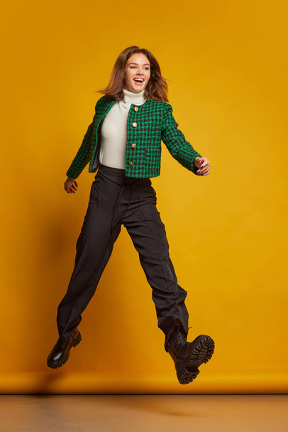 Portrait of young beautiful woman posing in stylish clothes, walking in a jump isolated over yellow studio background. Concept of youth culture, emotions, facial expression, fashion. Copy space for ad - Φωτογραφία, εικόνα