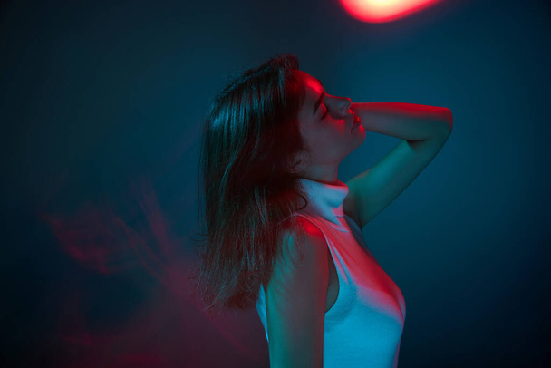 Lifestyle. Portrait of young beautiful woman posing, dancing isolated over blue studio background in red neon light. Concept of youth culture, emotions, facial expression, fashion. Copy space for ad - Photo, image