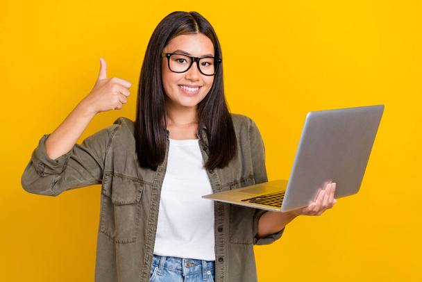 Portrait of positive optimistic woman with long hairdo wear khaki shirt show like good job hold laptop isolated on yellow color background. - Foto, immagini