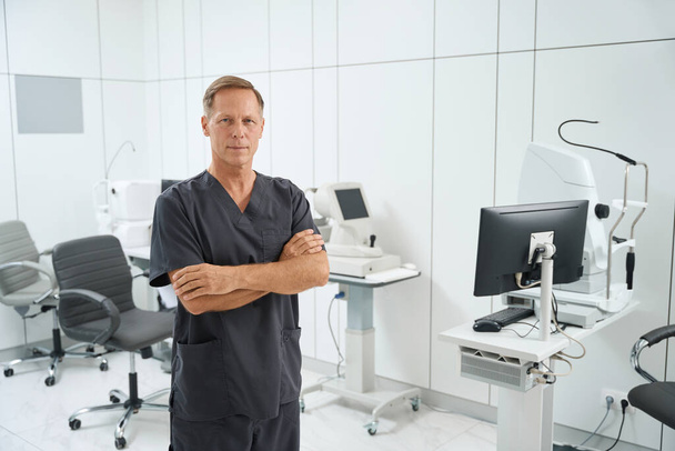 Portret lekarza w średnim wieku w garniturze medycznym stojącego w biurze ze sprzętem medycznym i patrzącego w kamerę - Zdjęcie, obraz