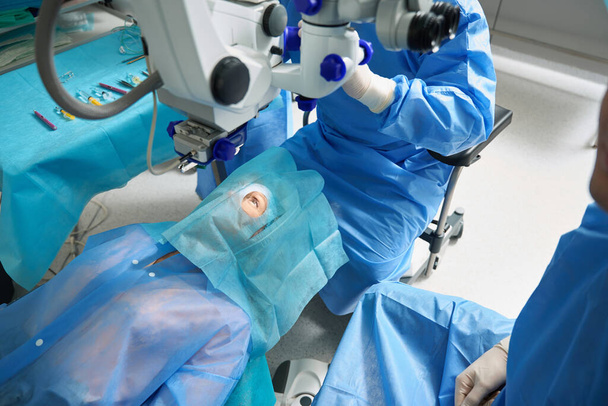 Γυναίκα με προστατευτική στολή ξαπλωμένη στο χειρουργικό τραπέζι και κοιτάζοντας ψηλά, ετοιμάζεται για χειρουργική επέμβαση στο μάτι - Φωτογραφία, εικόνα