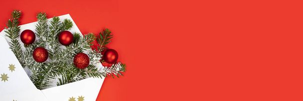 Christmas banner. White envelope with spruce branches and red Christmas tree balls on red background. Flat lay, place for text. - Photo, image