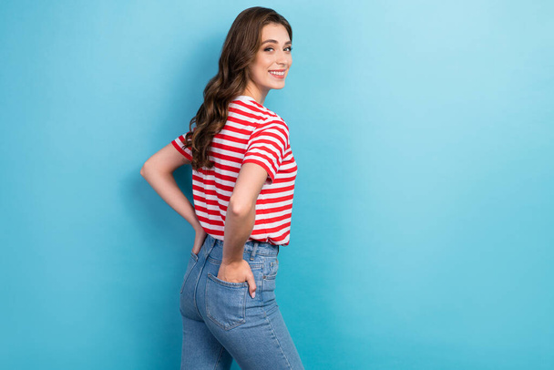 Photo of young adorable gorgeous nice pretty cute lady wear red striped t-shirt hands pockets looking back empty space isolated on blue color background. - Fotoğraf, Görsel