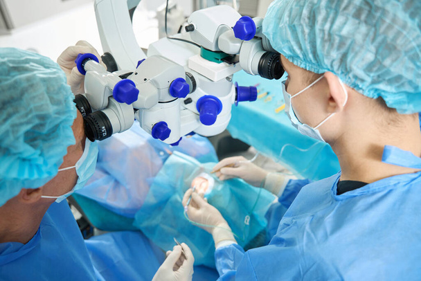 Korumacı, maskeli ve şapkalı cerrahlar hastayı mikroskop altında inceliyor. - Fotoğraf, Görsel