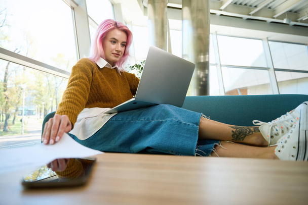 Vrouw zittend op de bank in coworking, kijkend op de laptop en het nemen van vel papier van de tafel - Foto, afbeelding