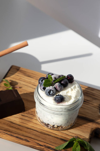 Jogurt w szklanym słoiku z granolą, czekoladą, jagodami i miodem na drewnianej desce na białym tle z liśćmi mięty. Widok z góry - Zdjęcie, obraz