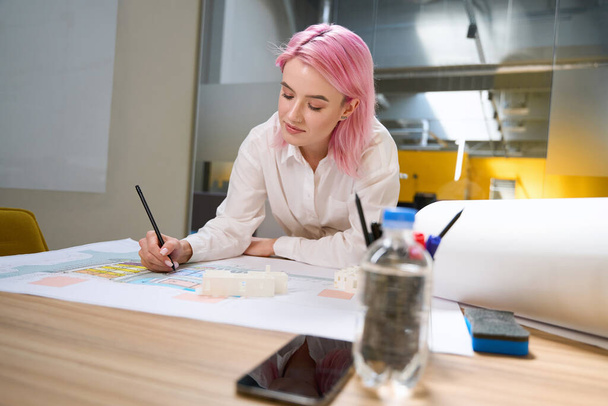 Νεαρός αρχιτέκτονας με ροζ μαλλιά κάθεται δίπλα στο τραπέζι, ενώ εργάζονται σε αρχιτεκτονικά έργα σε συνεργασία - Φωτογραφία, εικόνα