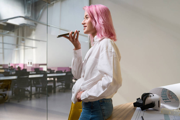Woman with pink hair holding phone, speaking with client in coworking, looking ahead and smiling - Valokuva, kuva