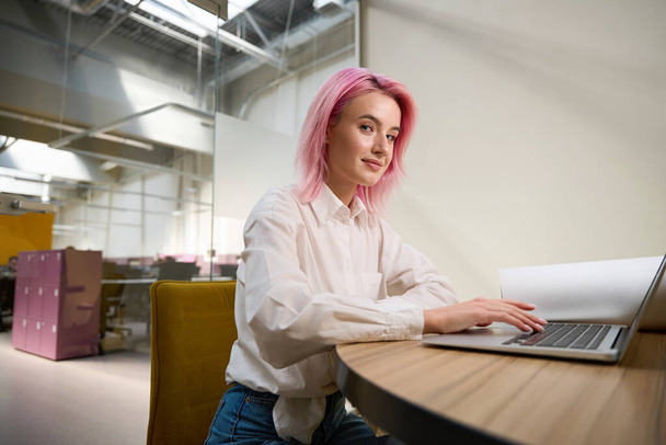 Pretty lady with pink hair sitting by the table with laptop in coworking and looking at the camera - Foto, Imagem