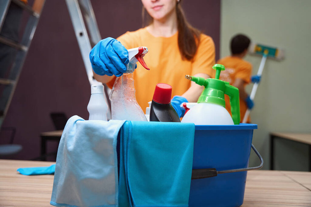 Обрезанное фото приятной юной леди, достающей дезинфицирующий спрей из коробки с помощью инструментов для уборки - Фото, изображение