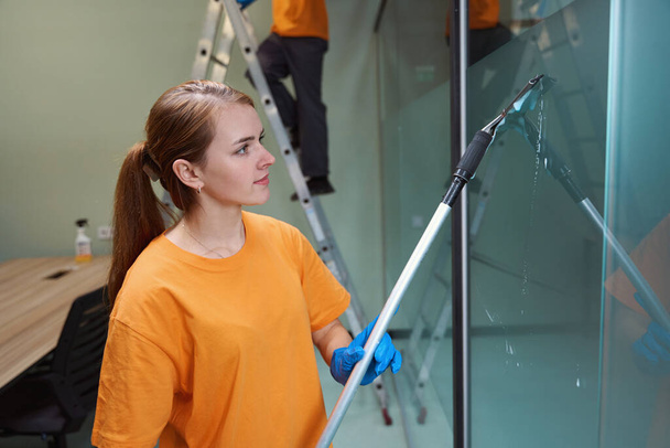 Mujer joven concentrada usando herramientas de limpieza de vidrio de interior mientras limpia la ventana - Foto, imagen