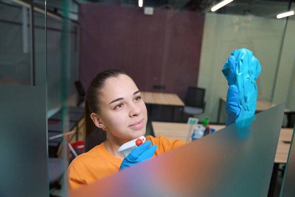 Mujer joven sonriente que usa guantes de goma mientras rocía productos químicos en la ventana y lo limpia - Foto, Imagen