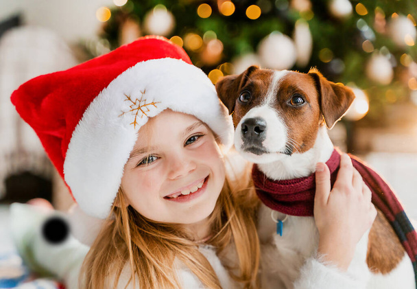 Petite fille mignonne profitant du temps de Noël avec son chien près du sapin de Noël en regardant la caméra - Photo, image