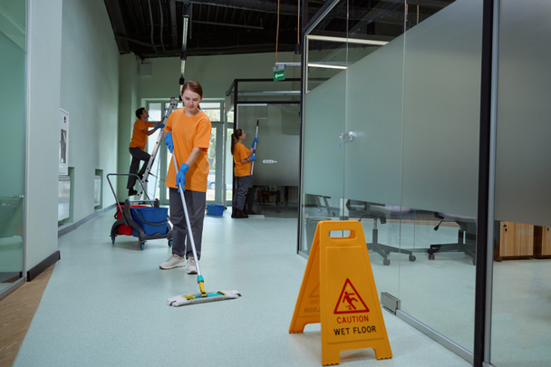 Óvatos női takarító nedves felmosóval a padlót a sárga figyelmeztető tábla mellett. - Fotó, kép