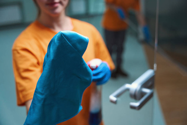 Καλλιεργημένη φωτογραφία προσώπου με πορτοκαλί πουκάμισο με χρήση υφάσματος και ψεκασμού χημικών κατά τον καθαρισμό της γυάλινης πόρτας - Φωτογραφία, εικόνα