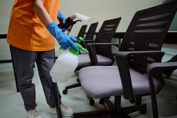 Обрезанное фото неизвестного человека в резиновых перчатках, распыляющего дезинфицирующее средство на стул - Фото, изображение