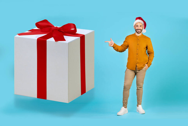 Um homem bonito em um chapéu de Natal em um fundo azul aponta o dedo para um grande presente branco com uma fita vermelha. Foto de alta qualidade - Foto, Imagem