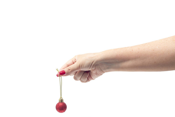 Una mano femminile, con le unghie dipinte di rosso, che tiene una palla di Natale rossa su uno sfondo bianco. - Foto, immagini