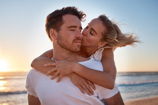 Love, couple and cheek kiss at beach on date, vacation or summer trip. Sunset, affection or romance of man and woman kissing at seashore, bonding and enjoying quality time together outdoors at coast - Фото, зображення