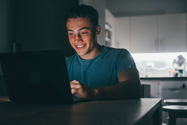 Een jonge tiener die 's avonds thuis studeert en werkt op de tafel met behulp van een laptop die lacht en plezier heeft in het huiswerk maken. Licht van het scherm op het gezicht. - Foto, afbeelding