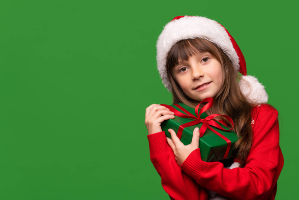 Маленька дівчинка тримає подарунок на зеленому ізольованому фоні. Дитина в капелюсі Санта-Клауса і теплий светр мають подарункову коробку. Фотографія високої якості - Фото, зображення