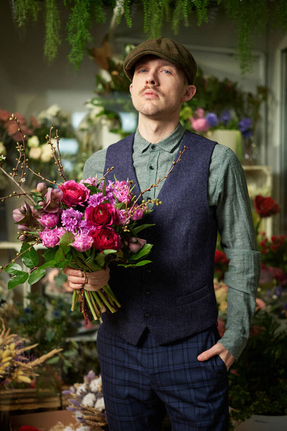 Homem caucasiano alegre atraente em boné de pico e roupas vintage de 20s segurando belo buquê de flores vermelhas e violetas. Conceito de Dia das Mães ou Dia dos Namorados. Imagem de alta qualidade - Foto, Imagem
