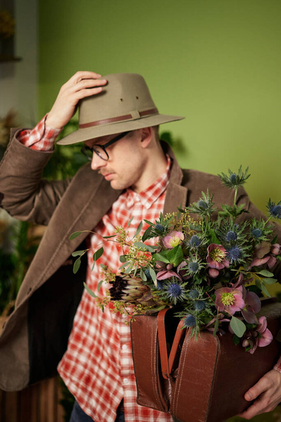 Vonzó vintage stílusú férfi személy szemüvegben, bársony kabát és aktatáska tartózkodik virágcsokor javító kalapját a virágboltban. Költő vagy író koncepció. Vvertikális kép - Fotó, kép