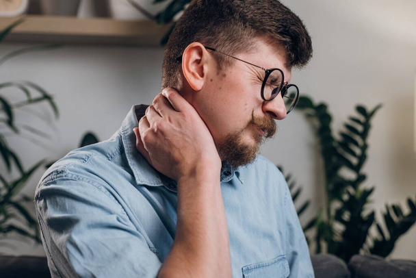 Young bearded man in eyeglasses studying until night at computer with neck pain. Close up of tired worker kneading neck, muscle recovery and massage. Neck pain, cervical osteochondrosis concept - Foto, Imagen