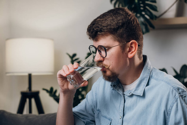 Handsome bearded male drinks water from glass. Useful habits and healthy lifestyle concept. Young man in eyeglasses works at home sitting on couch at home, looks at computer monitor and drinking water - Foto, imagen