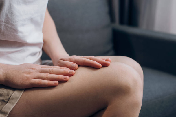 Close-up of female legs with fatigue and cramp. Legs pain concept. Unrecognizable unhealthy young woman with quadriceps pain, feels discomfort, does massage itself sitting on couch at home - Foto, afbeelding