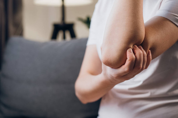 Close up of unhealthy young caucasian female touches elbow in morning and massages sitting on grey sofa at home, warming up injured hand after trauma - Valokuva, kuva