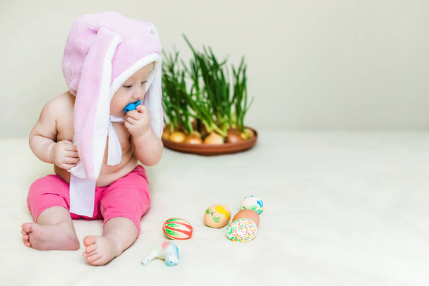 small beautiful baby sitter in rabbit hat. girl celebrates Easter and found painted decorative egg toy. love and religion concept - Photo, Image
