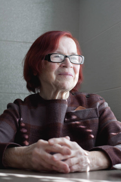 Porträt einer lächelnden älteren Frau mit roten Haaren, die eine Brille trägt. Die glückliche Großmutter schaut aus dem Fenster und lächelt. Alter - Foto, Bild