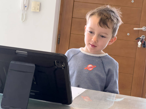 mignon caucasien garçon regardant attentivement tablette numérique sur table tout en étudiant en ligne à la maison - Photo, image