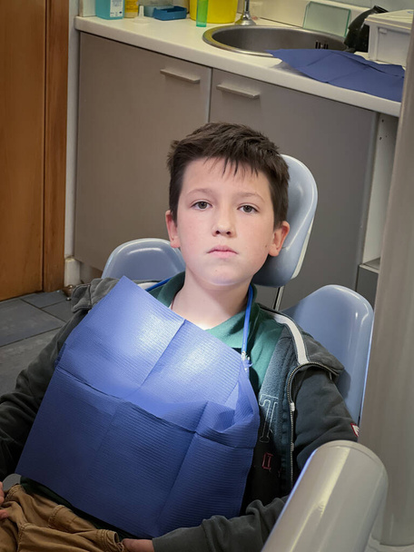 真剣な白人少年の肖像青のビブを着て歯科病院で歯医者の椅子に座って - 写真・画像