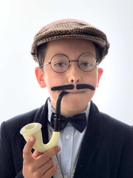 Πορτρέτο του αγοριού φορώντας κοστούμι με μουστάκι και επίπεδη καπέλο κρατώντας πίπα κάπνισμα και προσποιείται ότι είναι ντετέκτιβ σε λευκό φόντο - Φωτογραφία, εικόνα