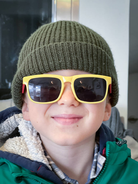 Retrato de cerca del lindo chico caucásico sonriendo y usando sombrero de punto y gafas de sol en casa - Foto, Imagen