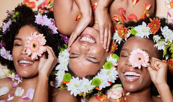 Top view, beauty and black women with flowers for skincare in studio on brown background. Makeup aesthetics, organic cosmetics and group, friends or female models with floral plants for healthy skin. - Foto, Imagem