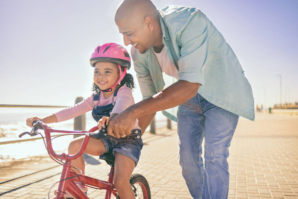 Black girl, bike and learning with dad, smile and park in sunshine, safety or happiness by sea. Father, kid and bicycle for summer training, childhood development and outdoor in street with helmet. - Φωτογραφία, εικόνα
